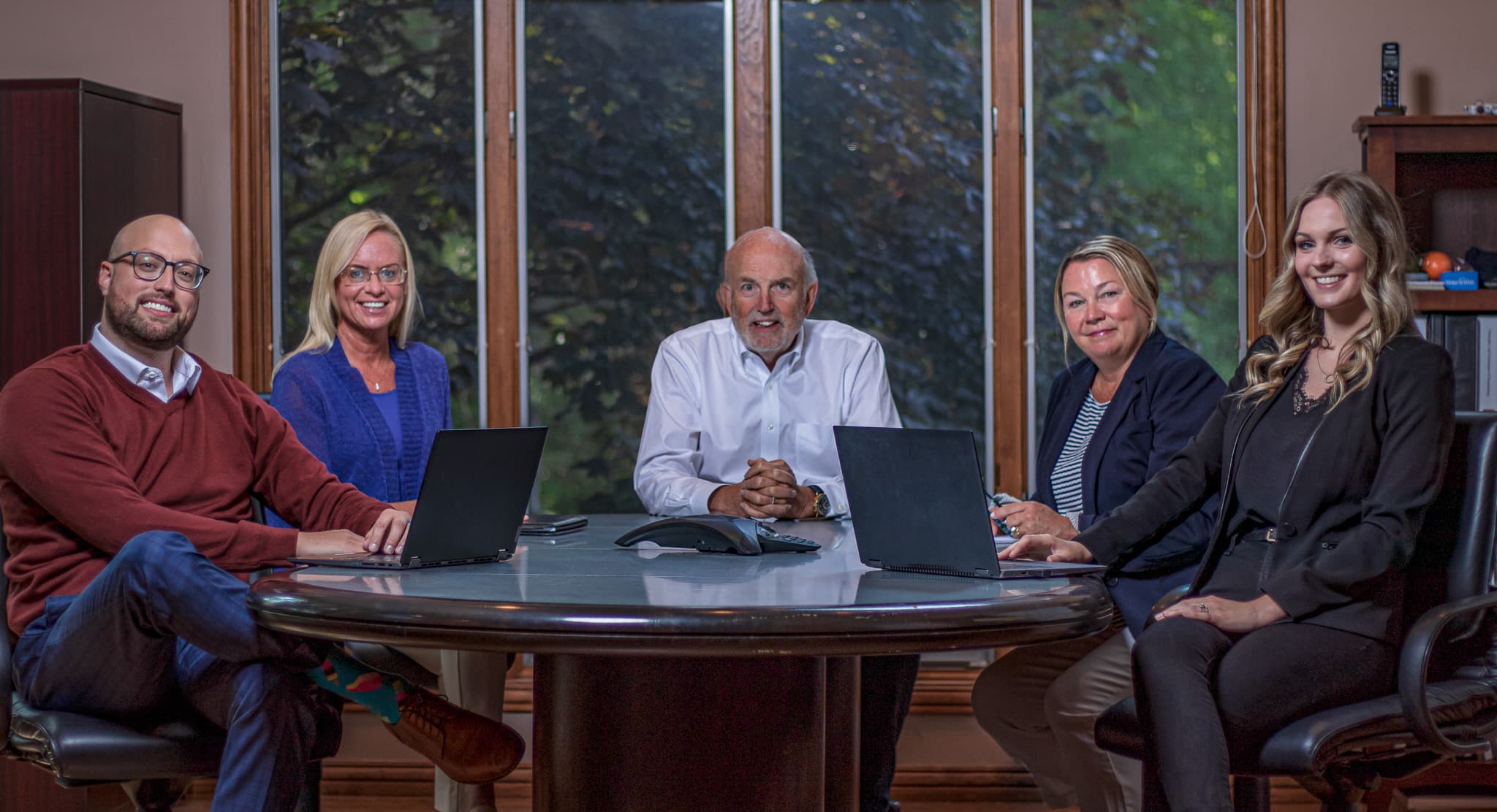 Five smiling business professionals seated around a conference table with laptops in an office.
