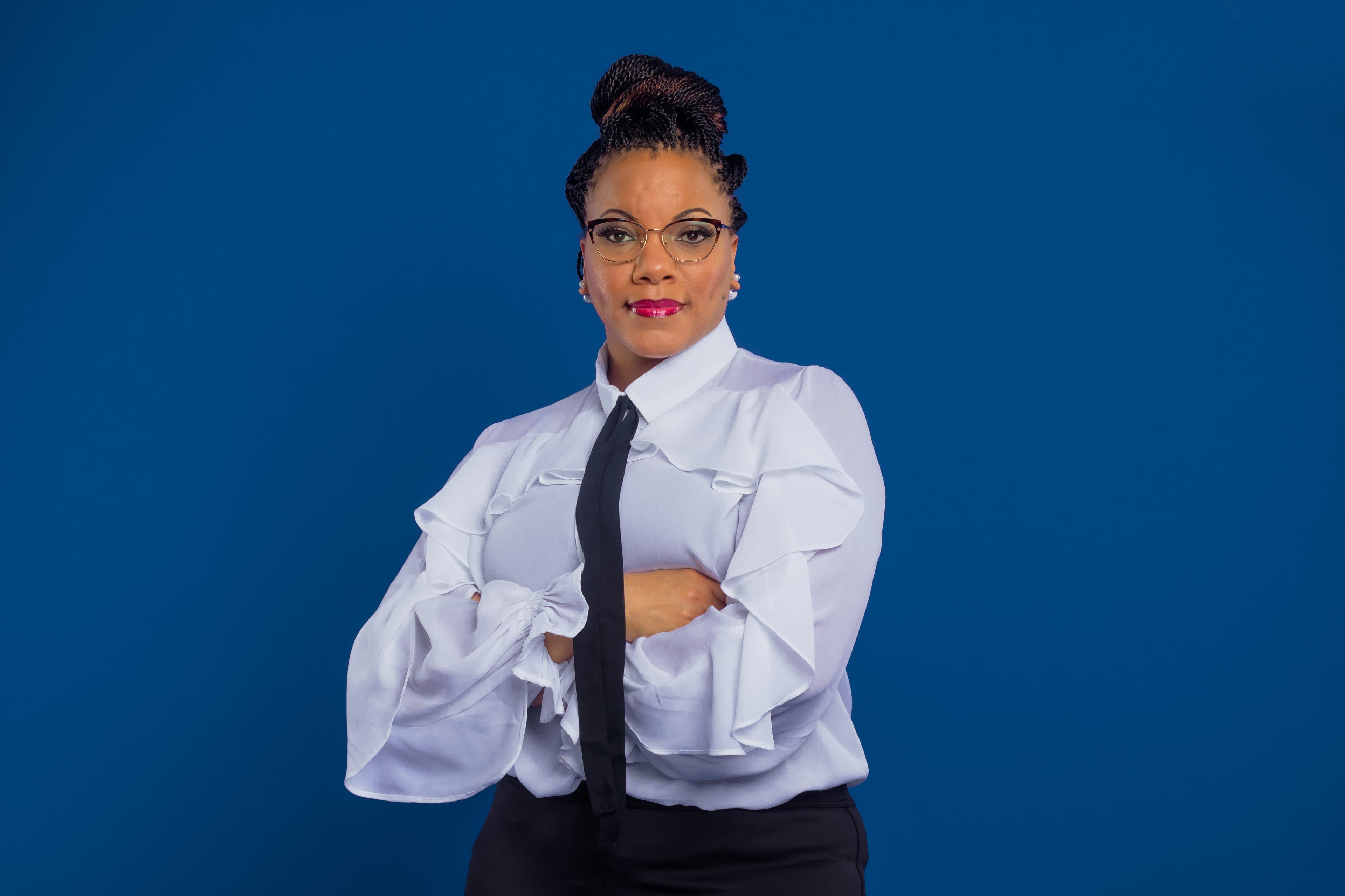 Black woman with braided bun and glasses, wearing a ruffled white blouse and black tie.