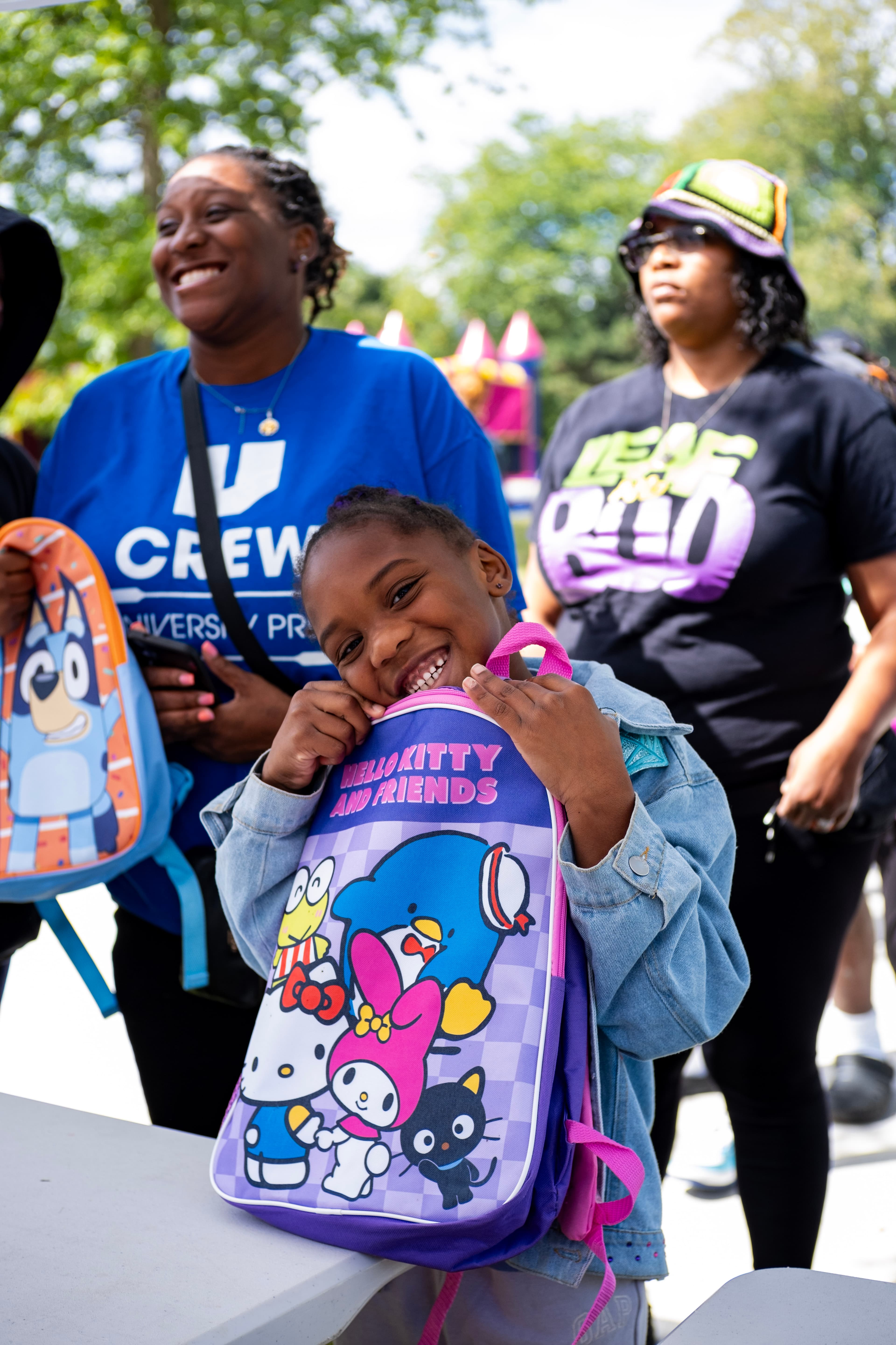 Smiling young girl in a denim jacket hugs a purple Hello Kitty backpack outdoors.