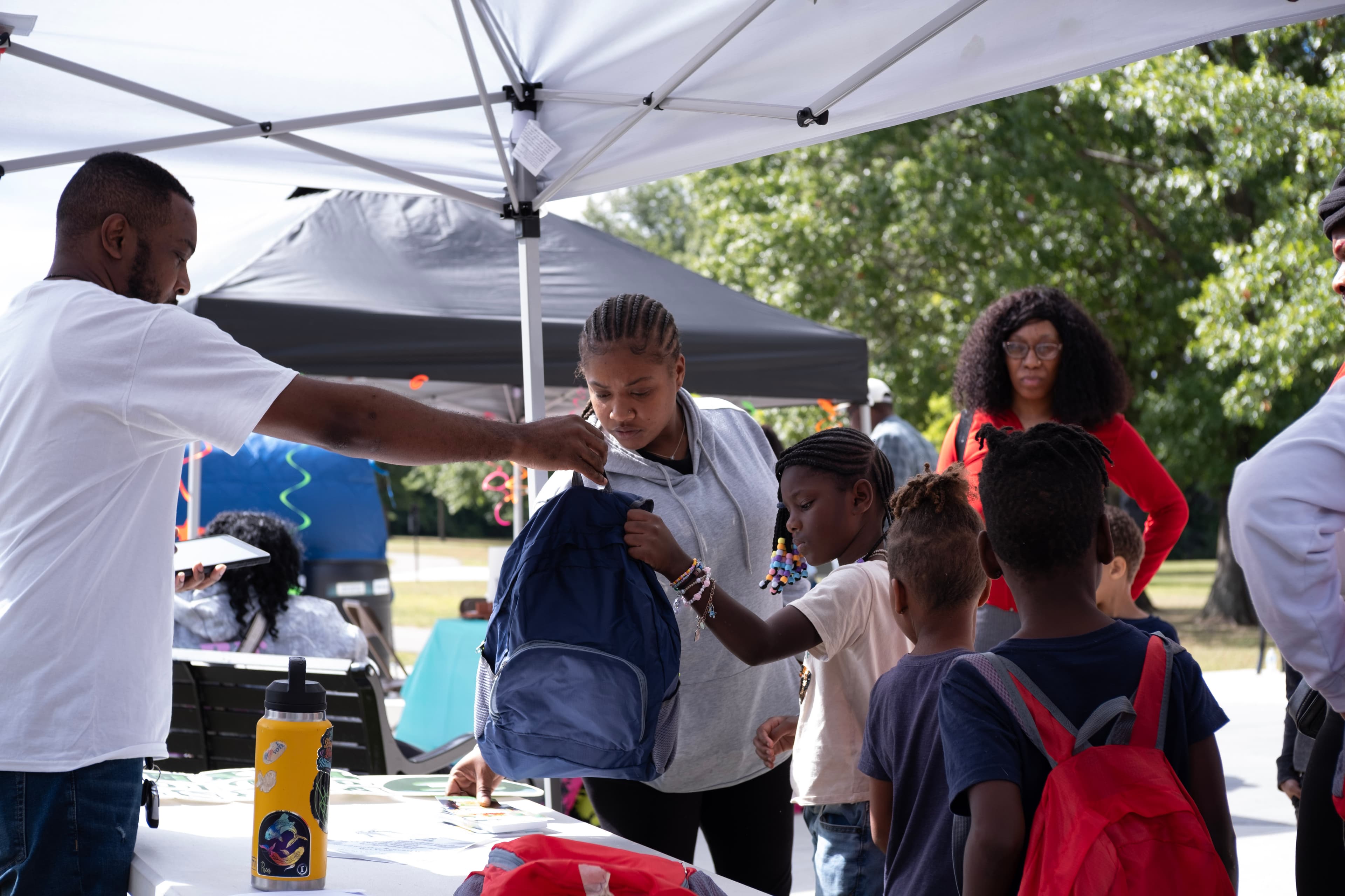 Man hands a blue backpack to a woman and child at a community giveaway event.