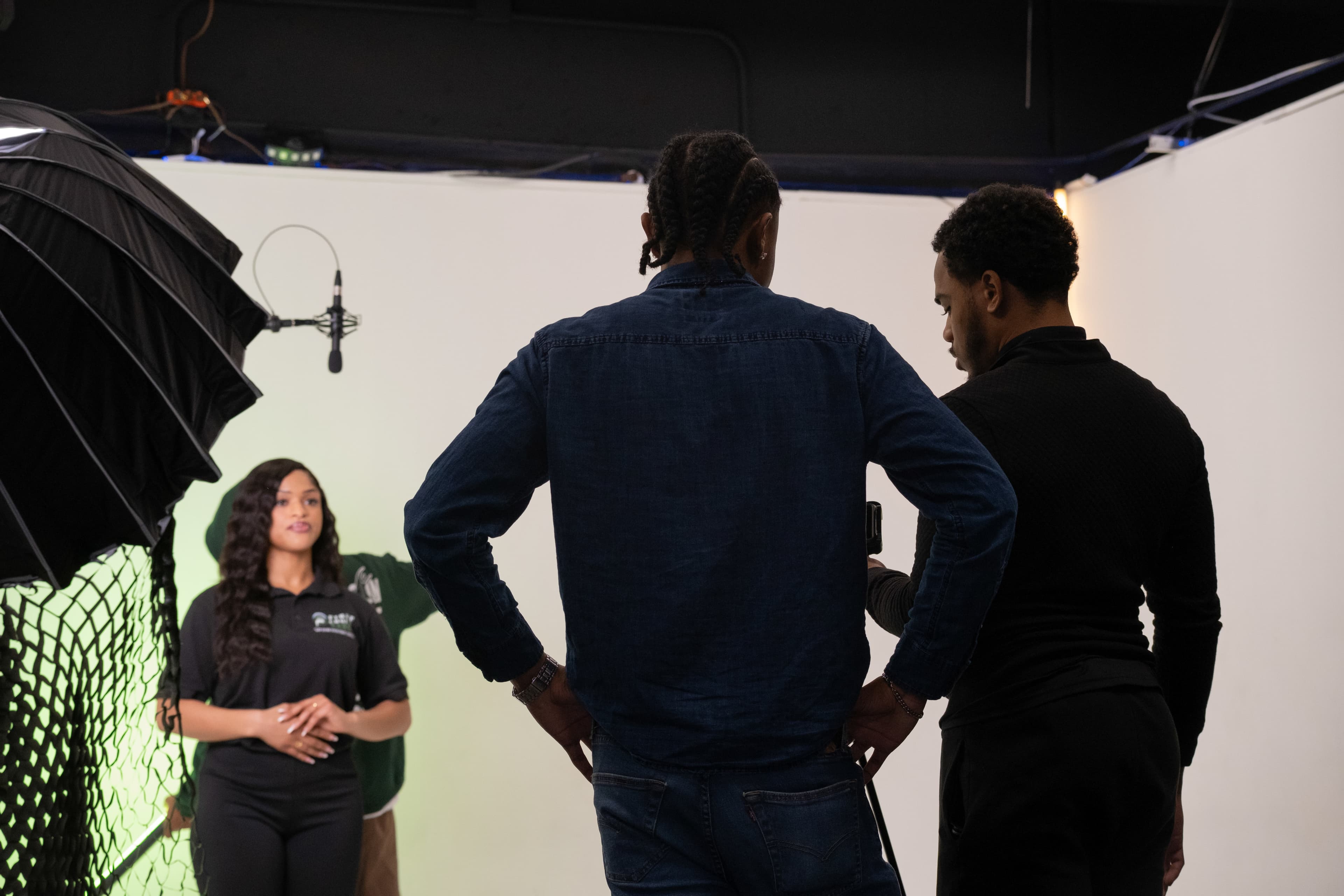 Woman posing in a studio while two men observe behind professional lighting and a microphone.