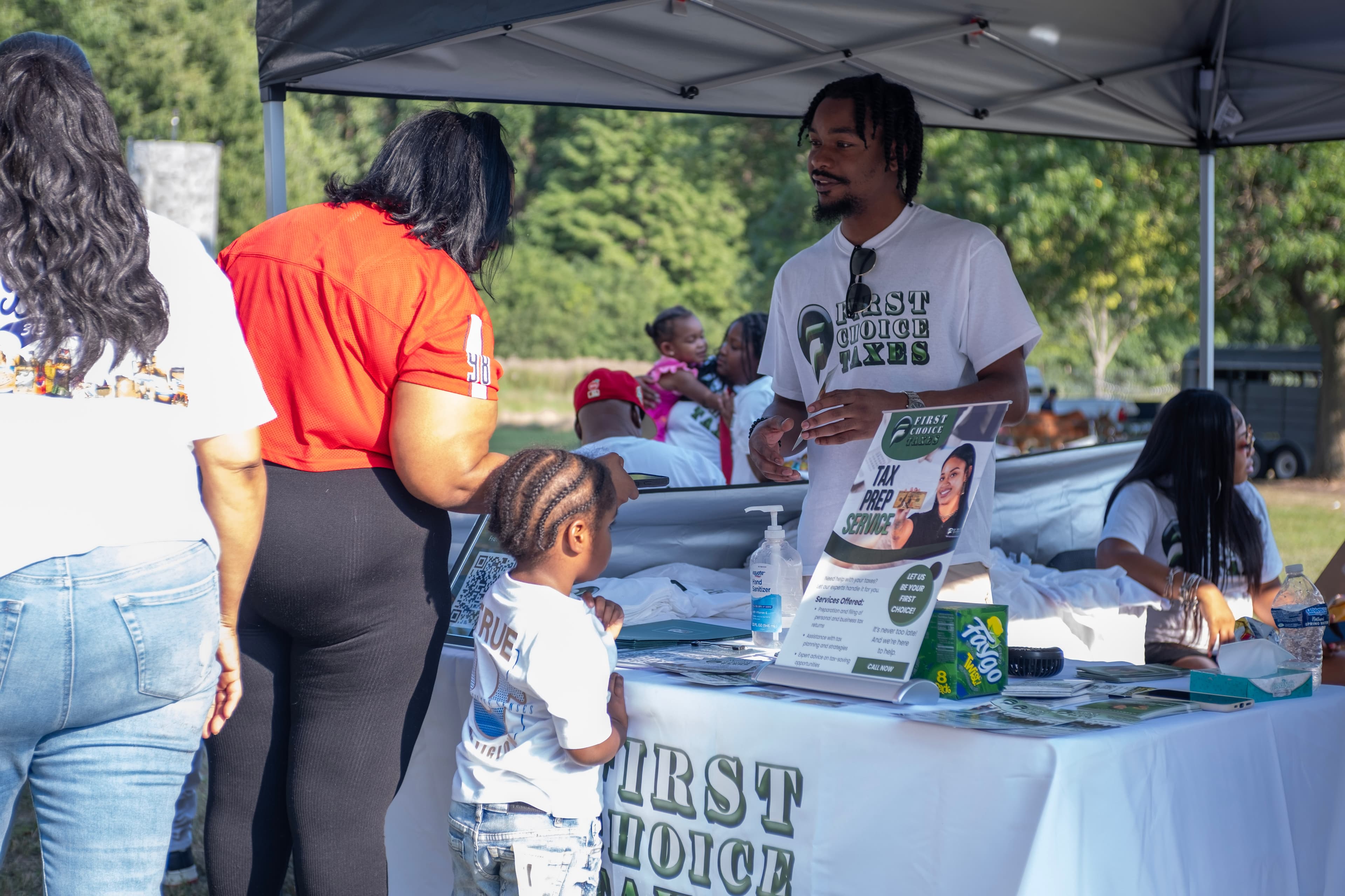 A man at a First Choice Taxes booth interacts with a woman and child outdoors.