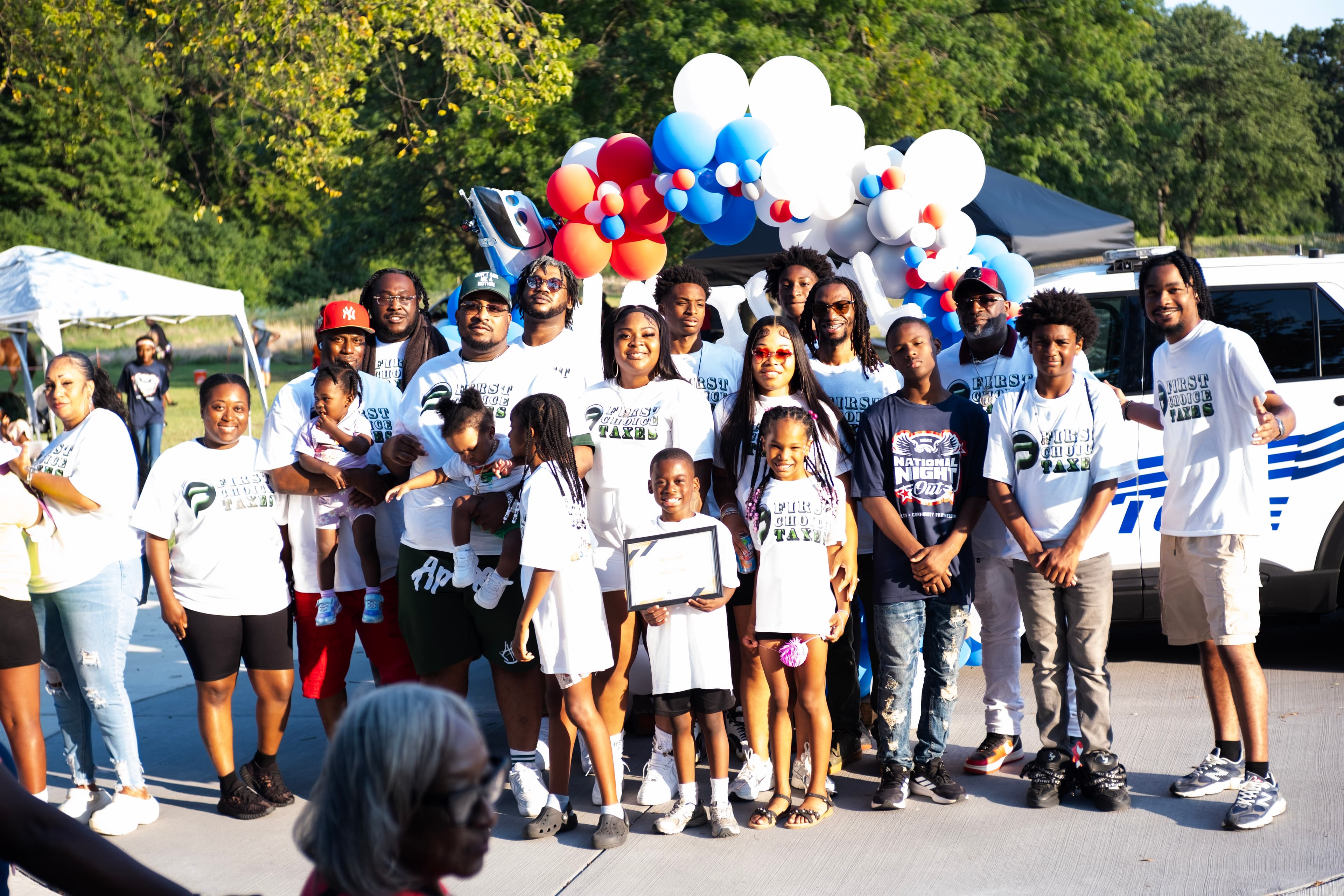 Group in First Choice Taxes shirts posing outdoors with red, white, and blue balloons.