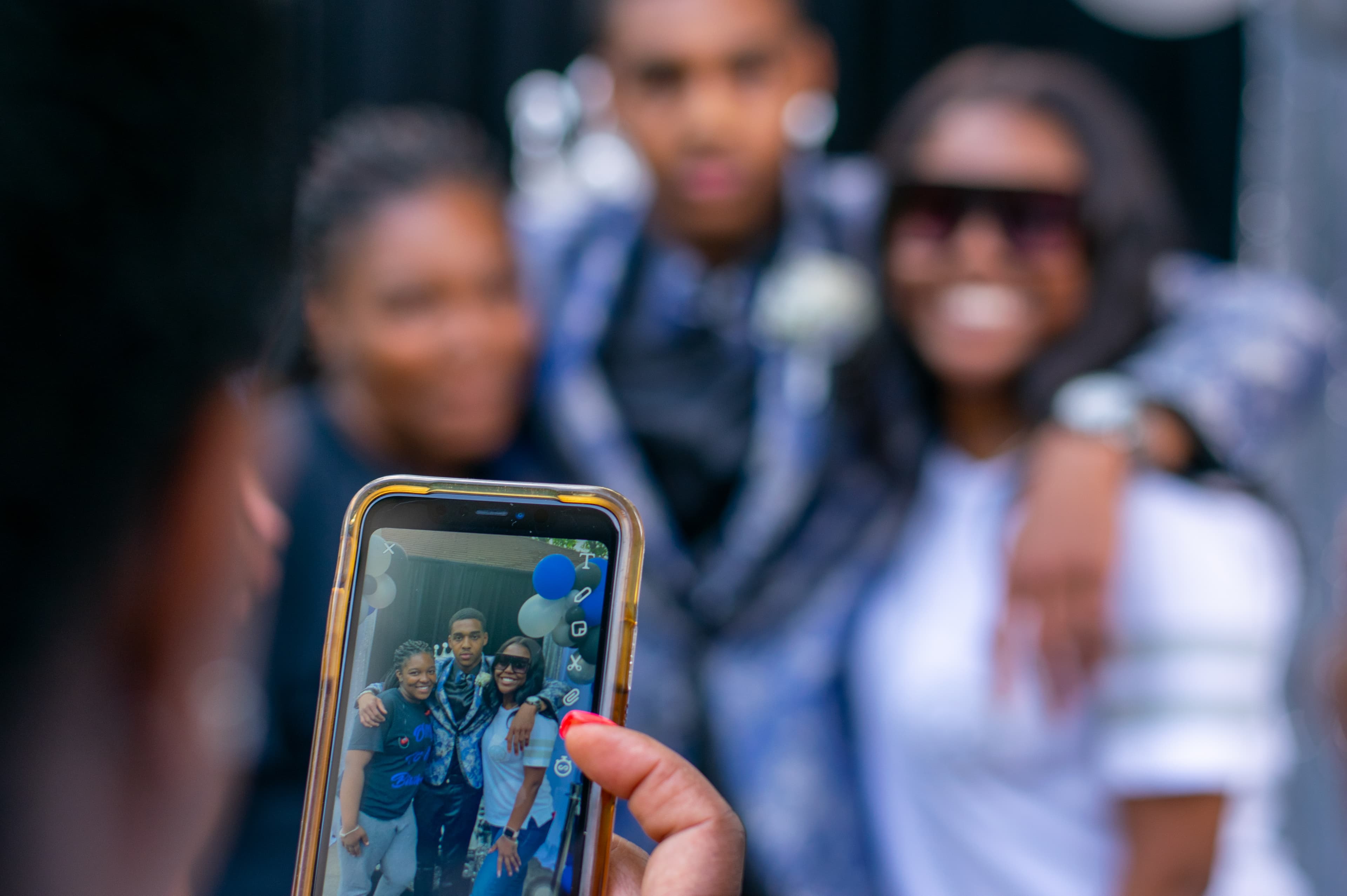Hand holding a smartphone to photograph three people, with the subjects blurred in background.