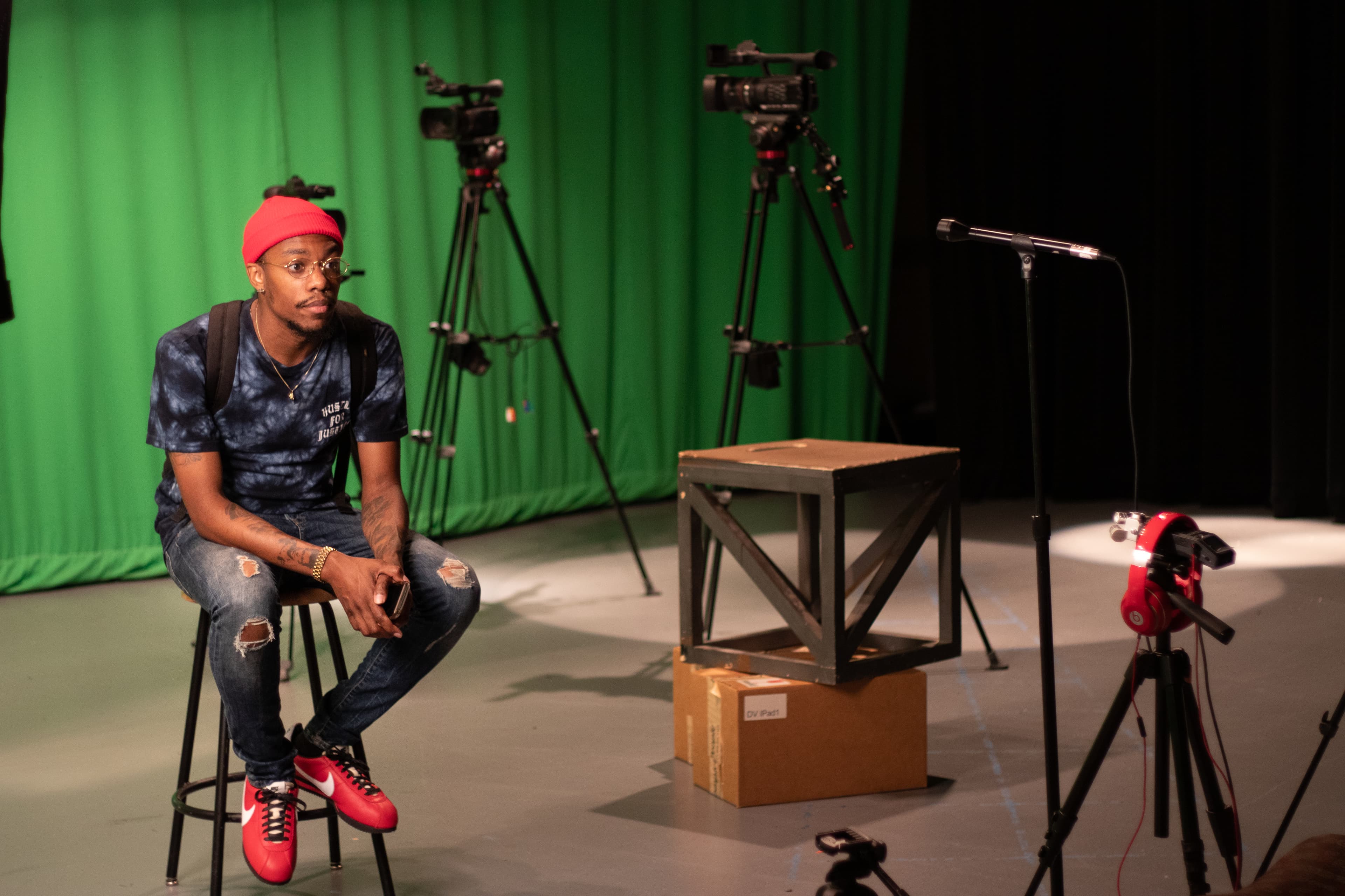 Man in red beanie and tie-dye shirt sits on stool in green screen studio.
