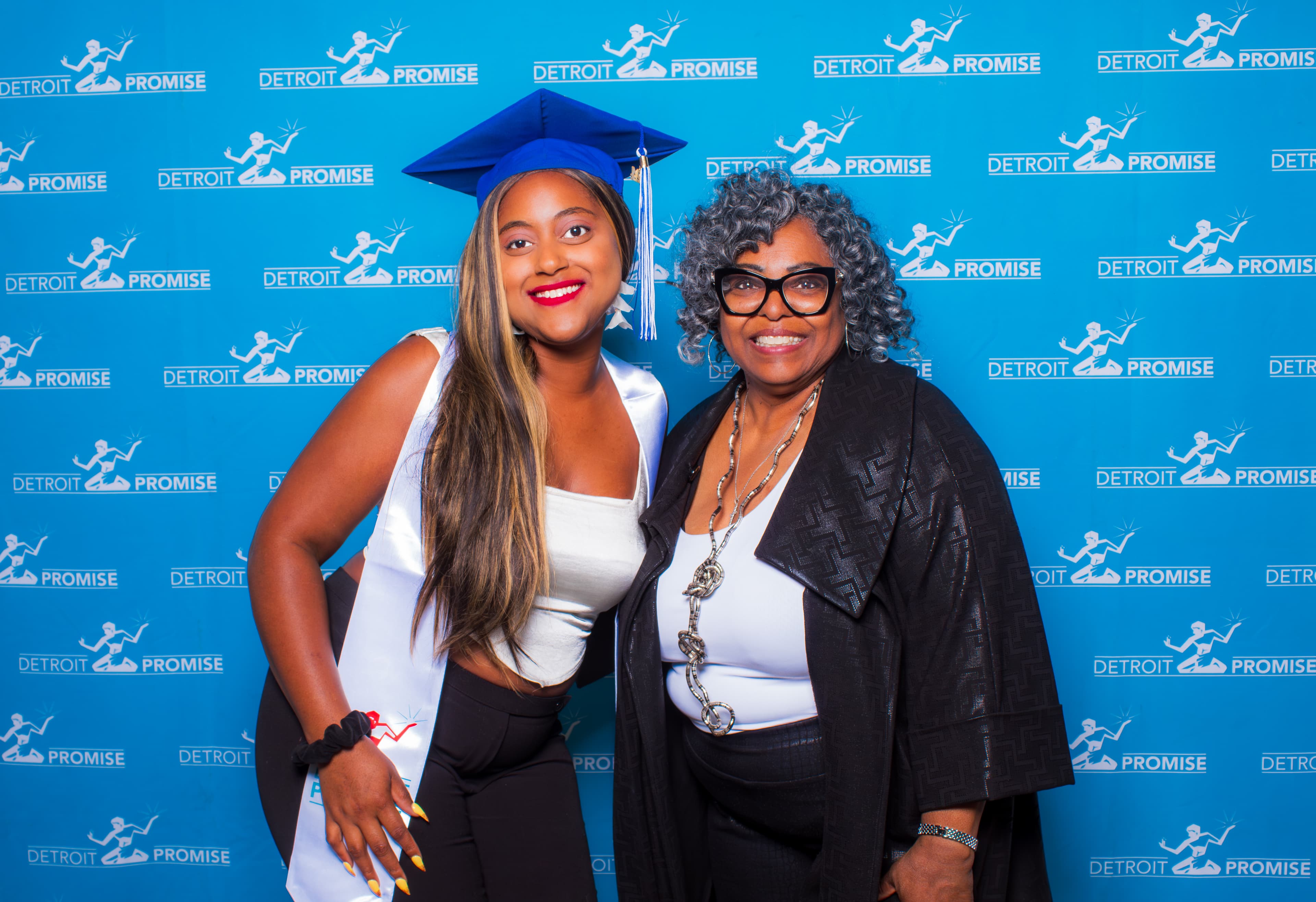 Graduate in blue cap poses with an older woman against a Detroit Promise backdrop.