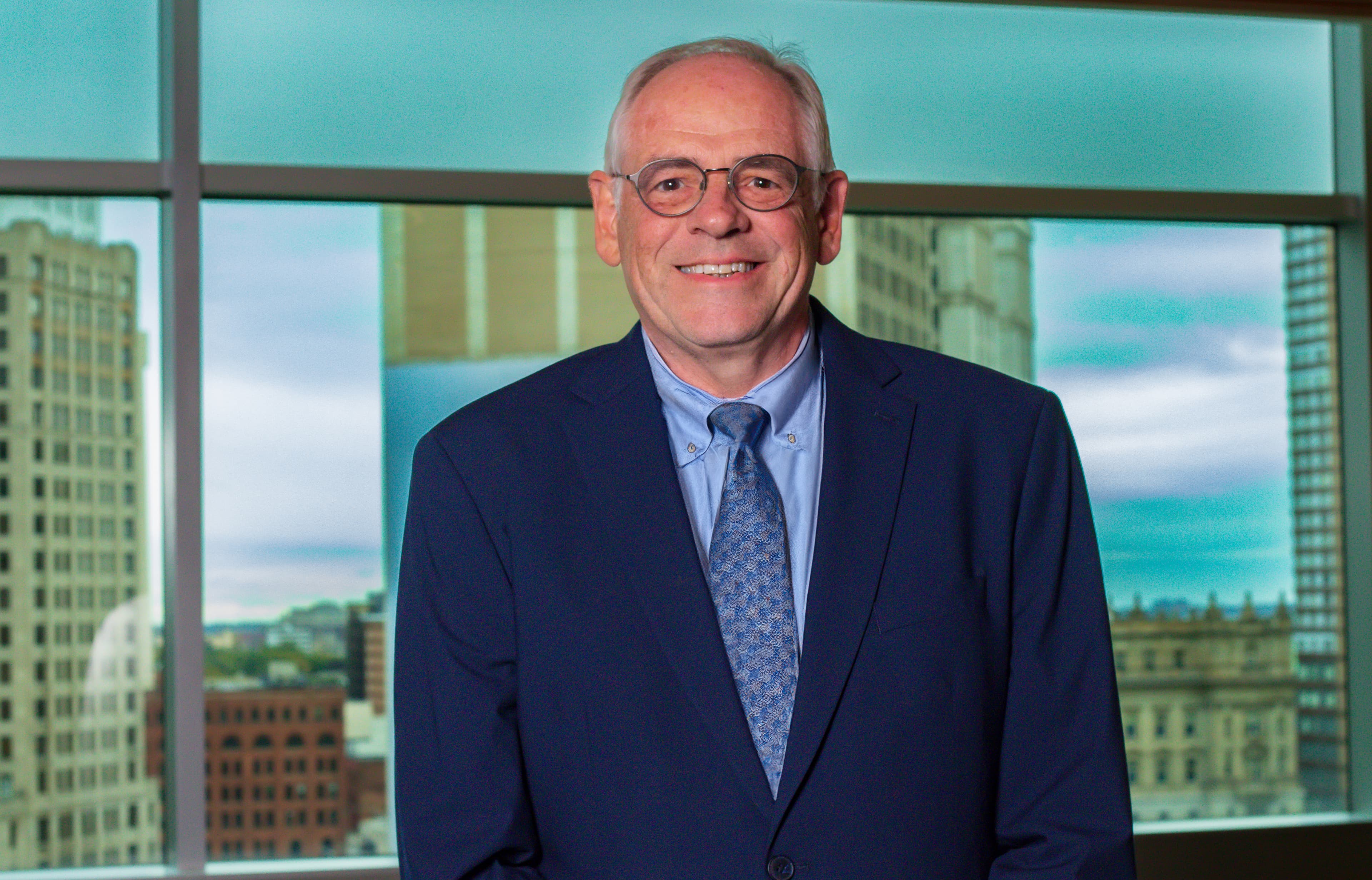 Smiling man with glasses wearing a navy suit and tie before a city skyline.