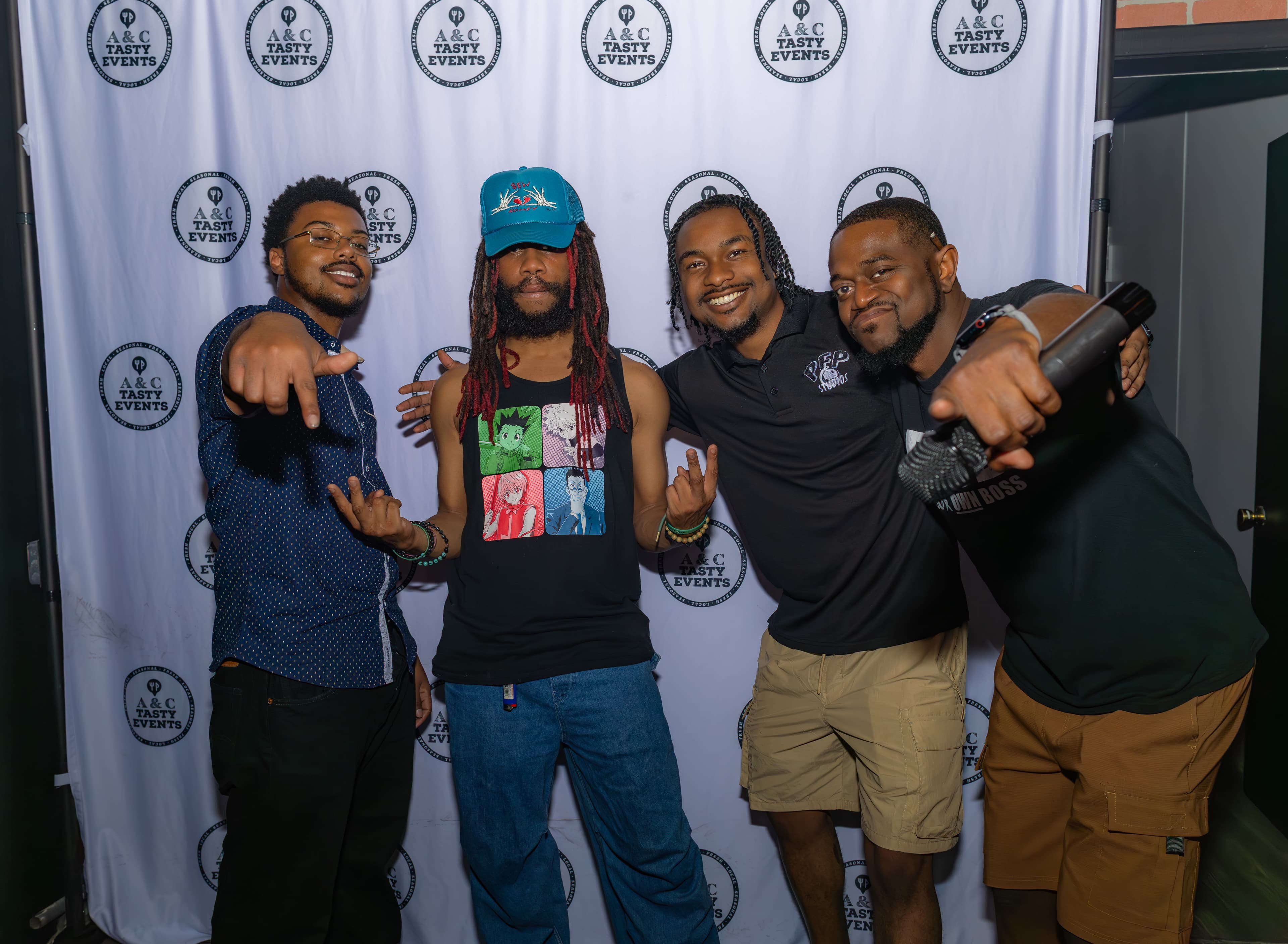 Four men, one holding a microphone, pose and gesture before a white event backdrop.
