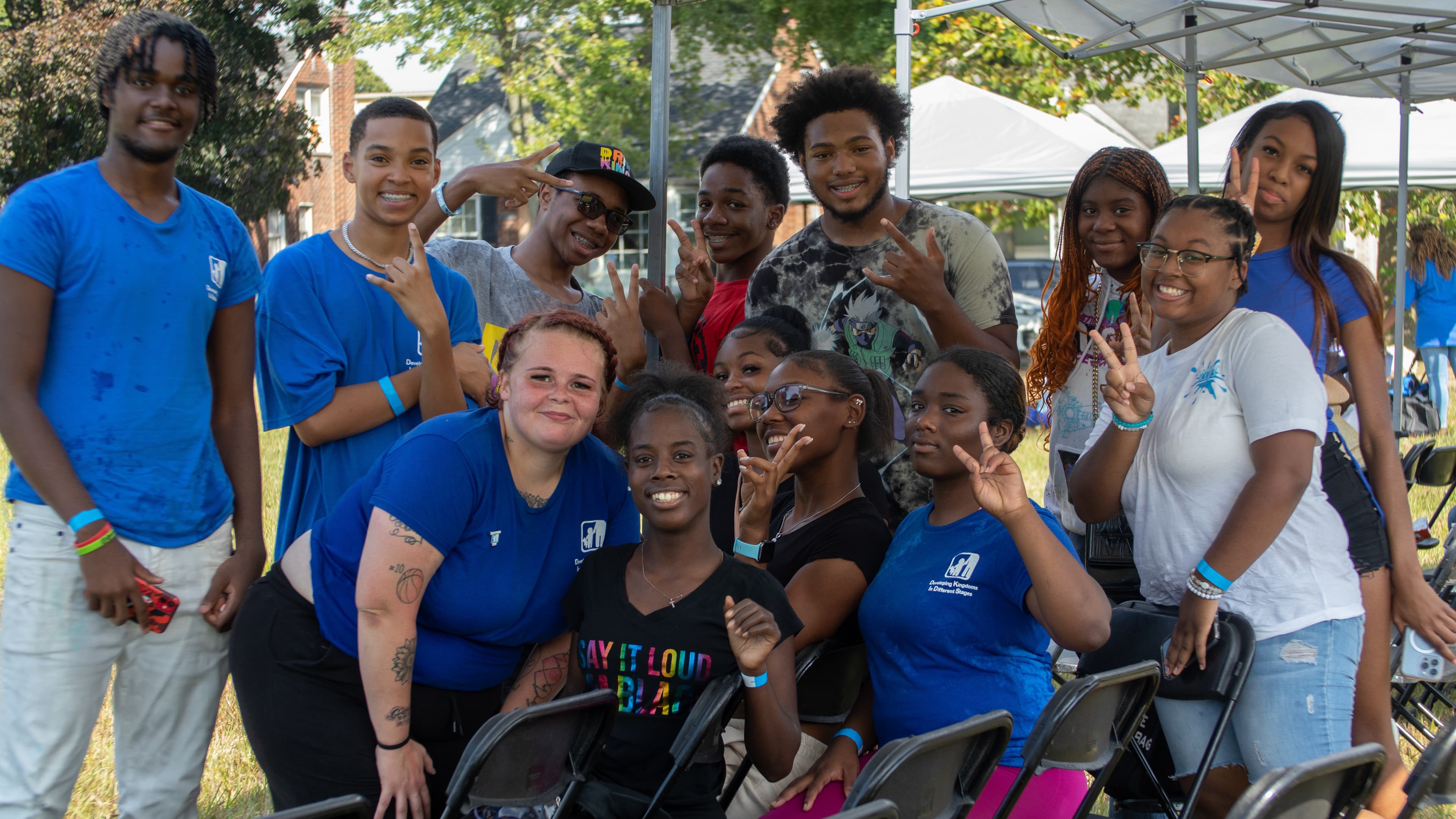 Diverse group of smiling young people posing outdoors and making hand signs under a canopy.