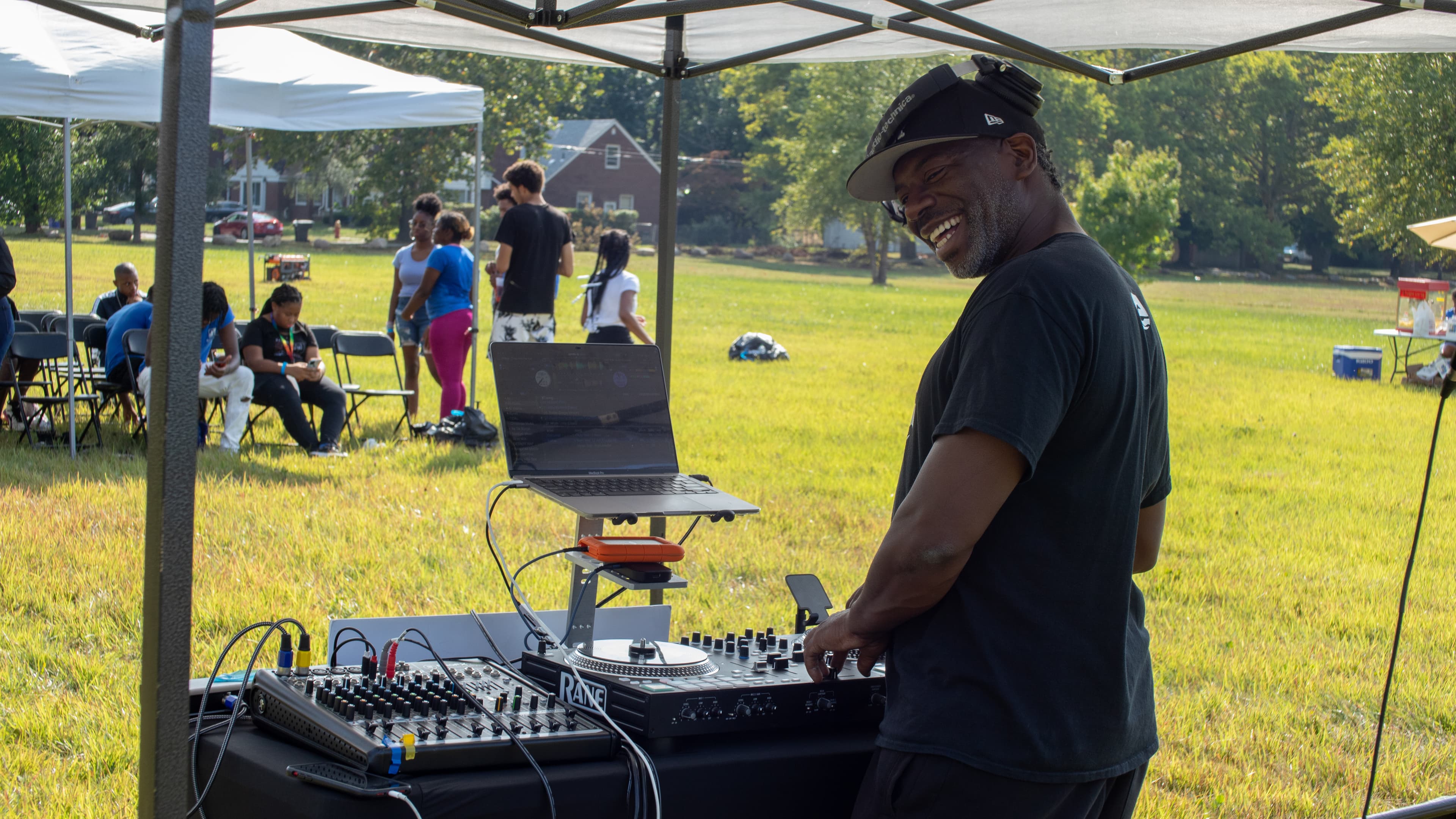 Smiling DJ operates a mixer and laptop under a canopy at a sunny outdoor event.