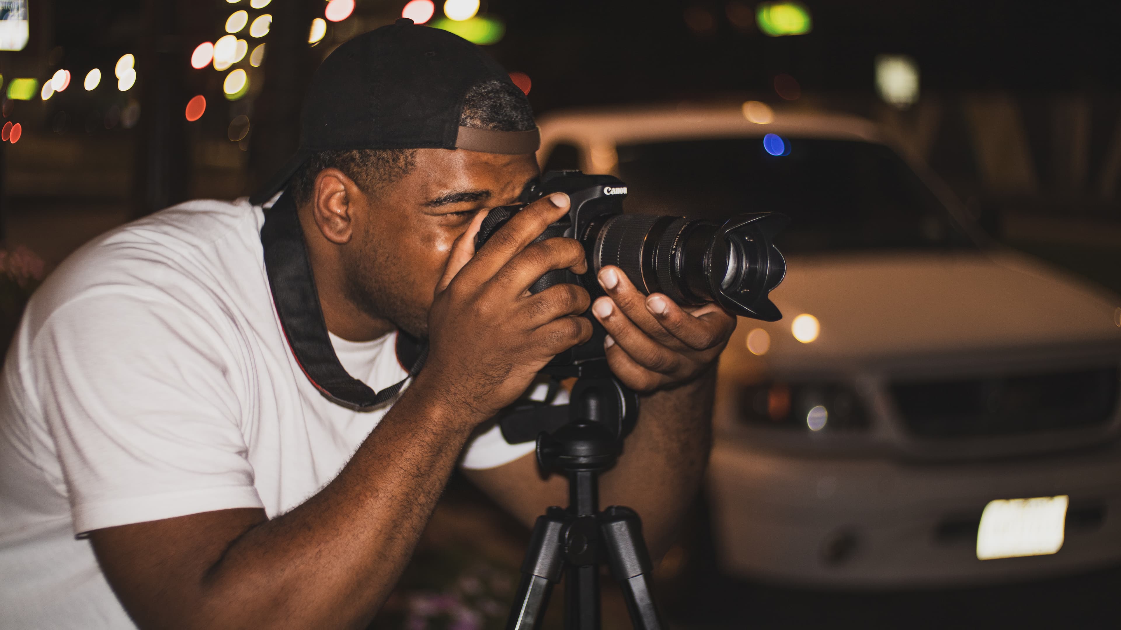 Photographer in a white shirt and black cap uses a tripod-mounted camera at night.