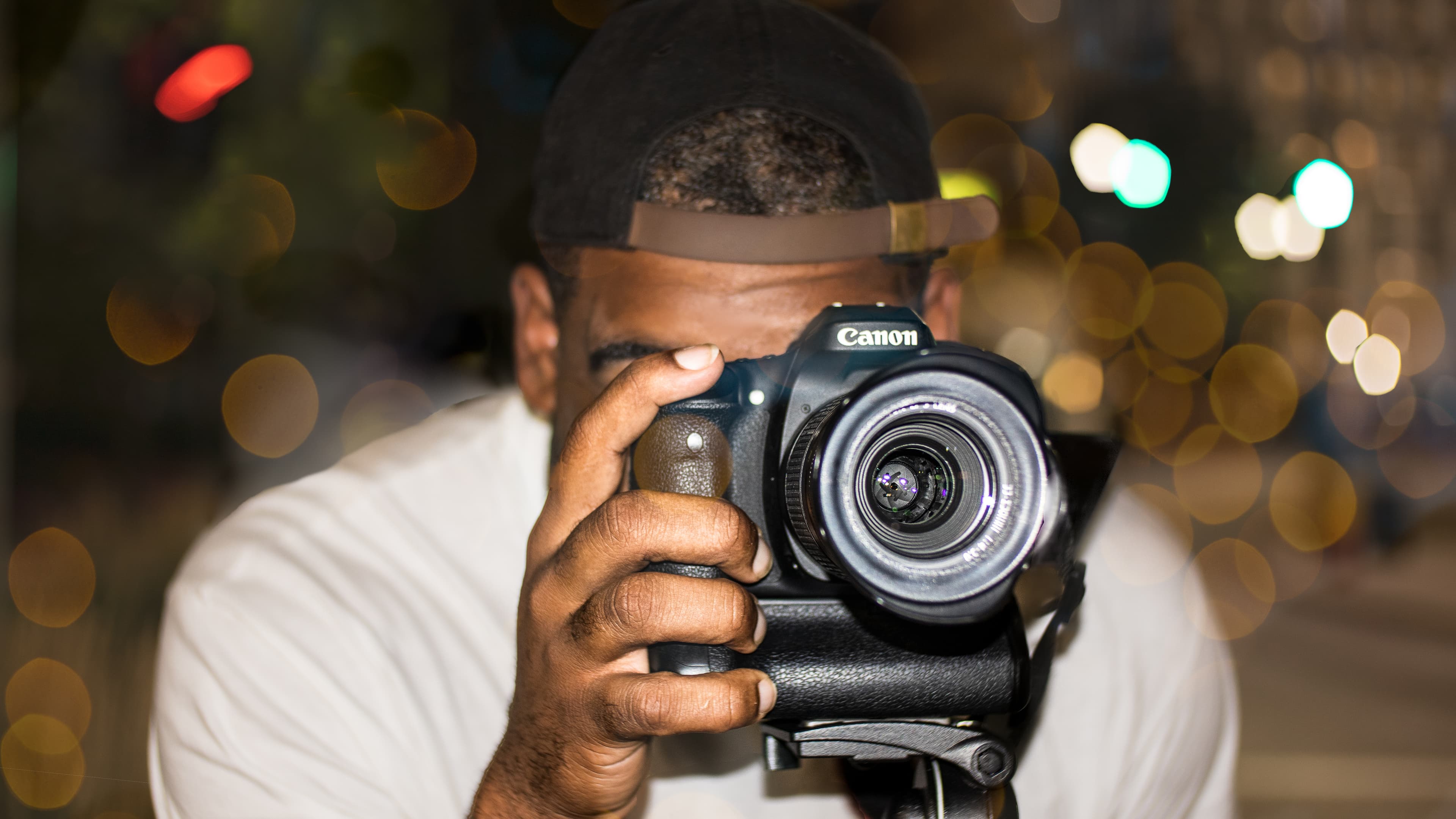 A photographer holds a Canon camera to his eye against blurry city lights at night.