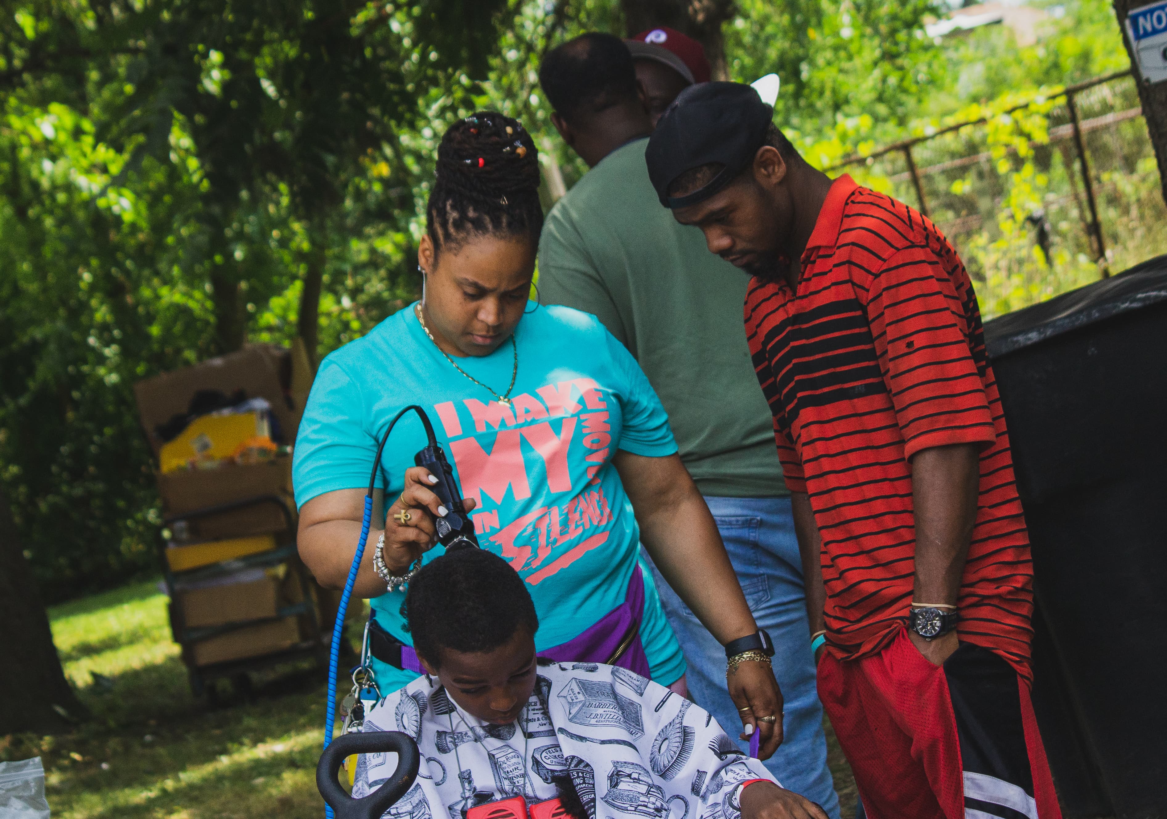Woman in turquoise shirt cuts a boy's hair with clippers outdoors while a man watches.