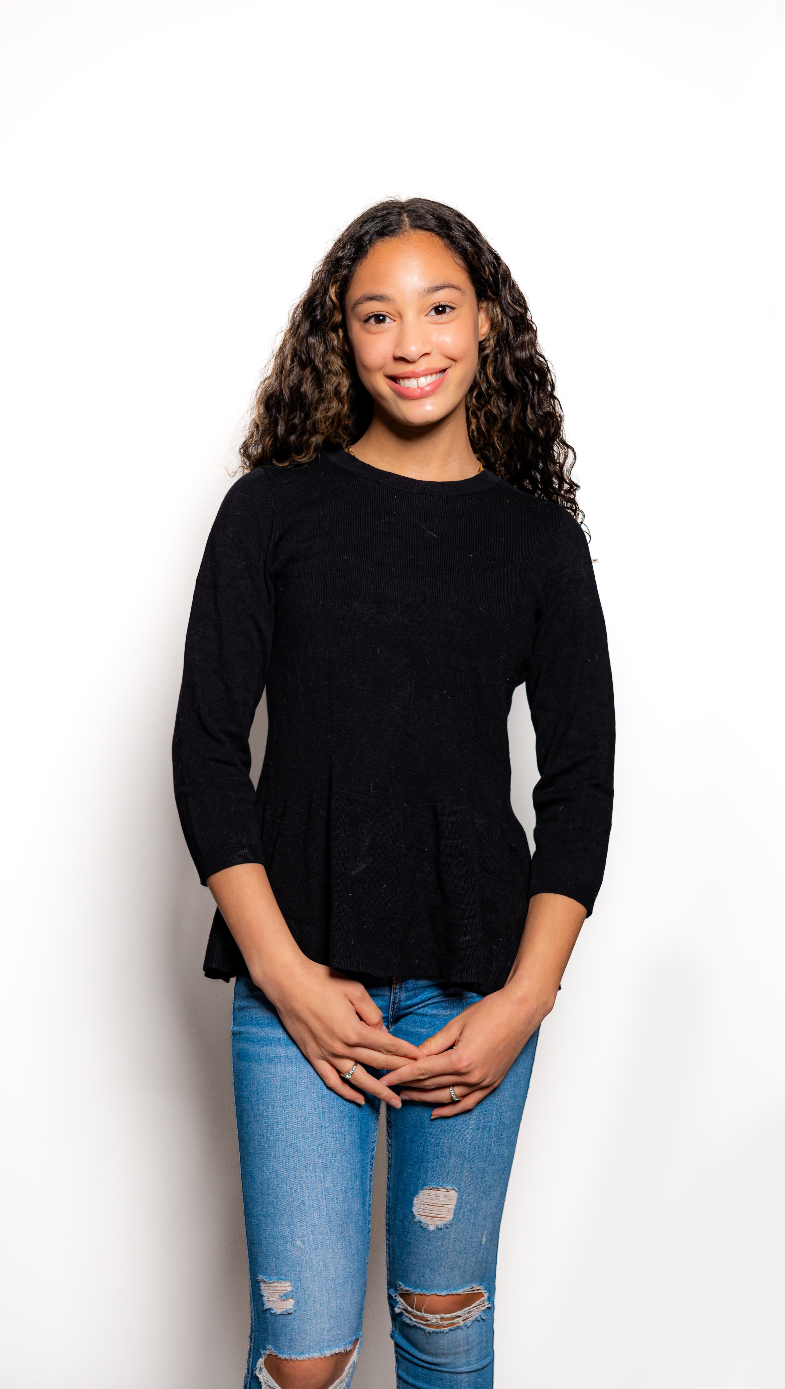 Smiling young woman with long curly hair wearing a black top and ripped blue jeans.