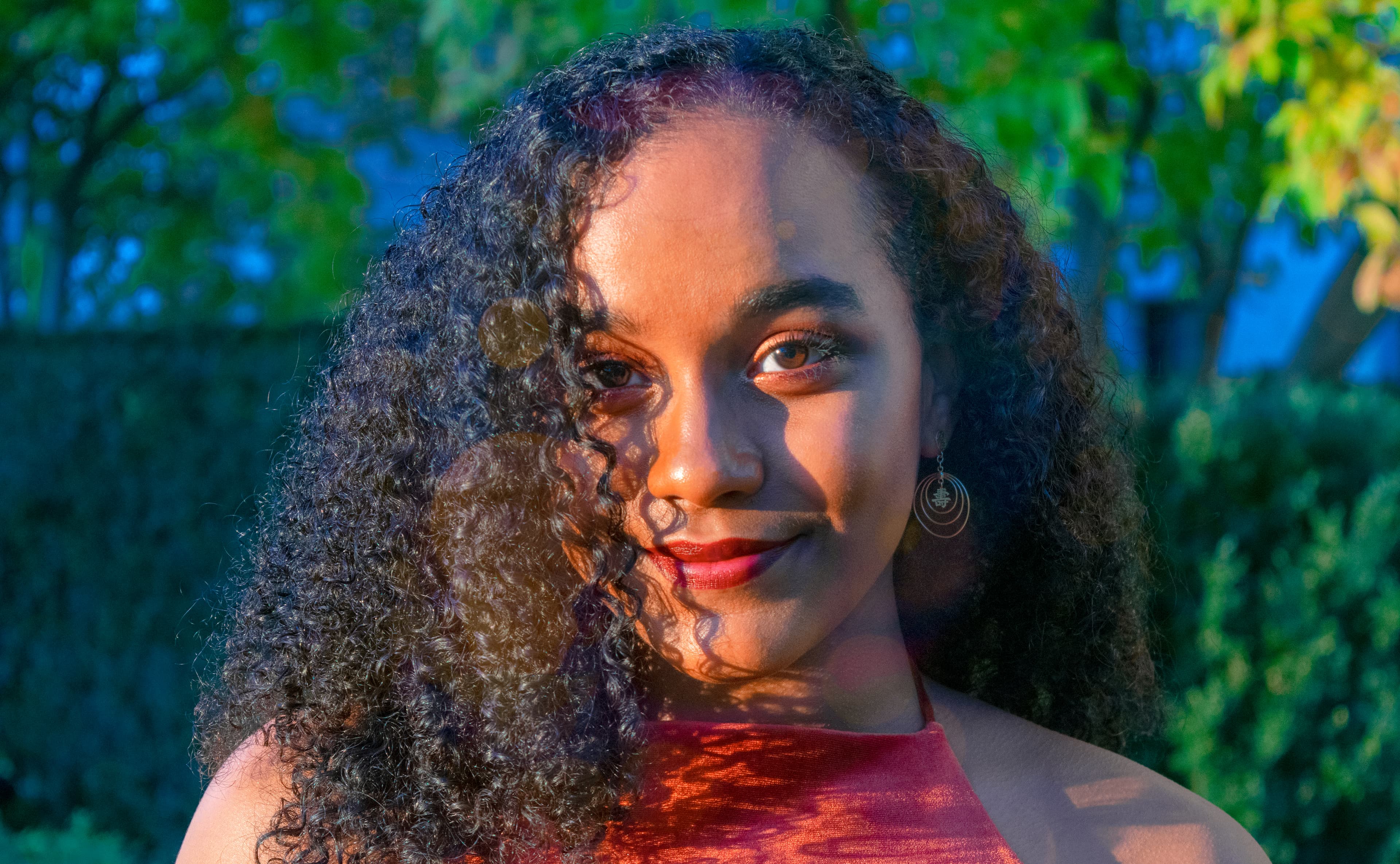 Woman with dark curly hair smiles as warm sunlight casts intricate shadows across her face.