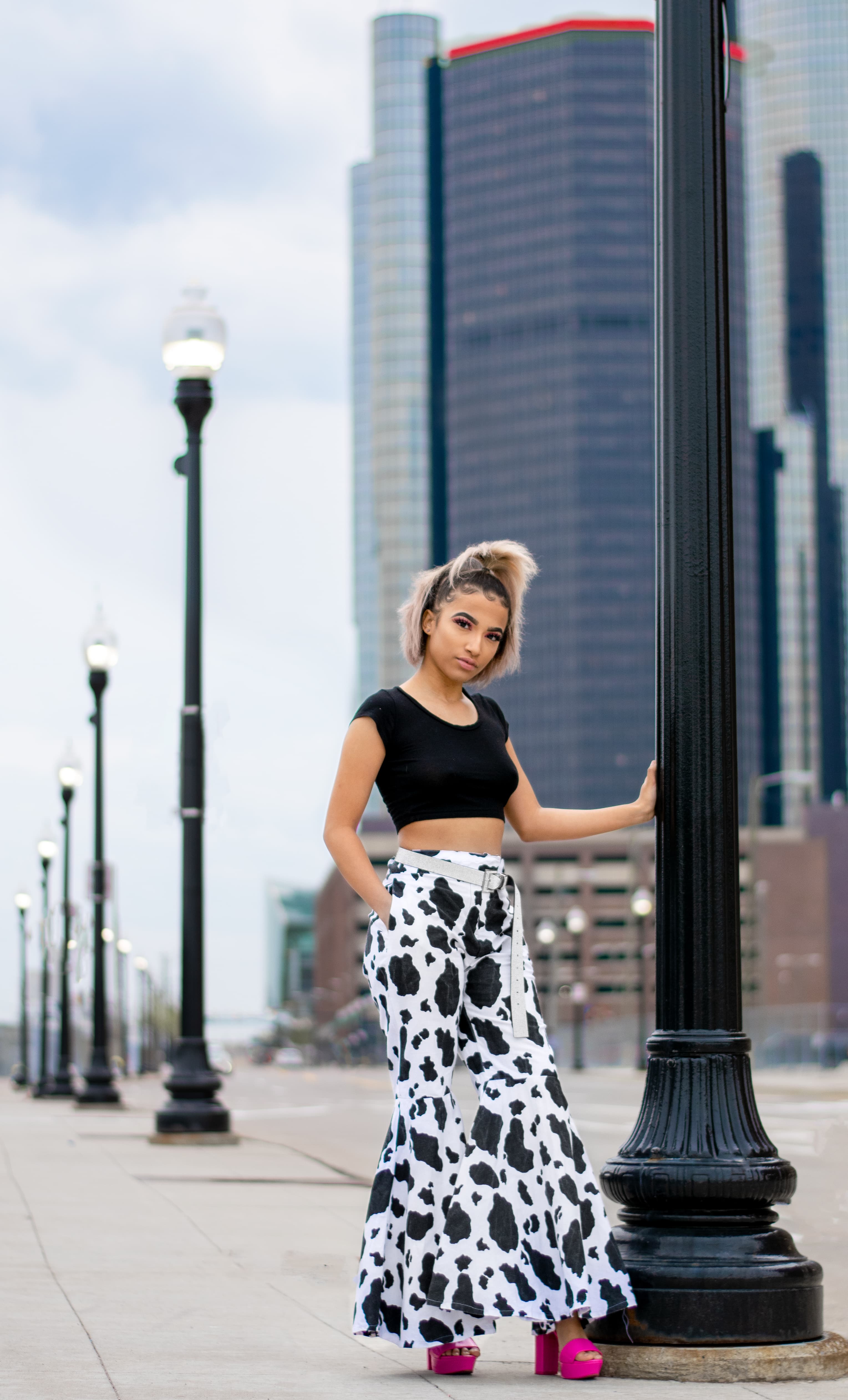 Woman in cow print flare pants and pink heels poses by a city lamp post.