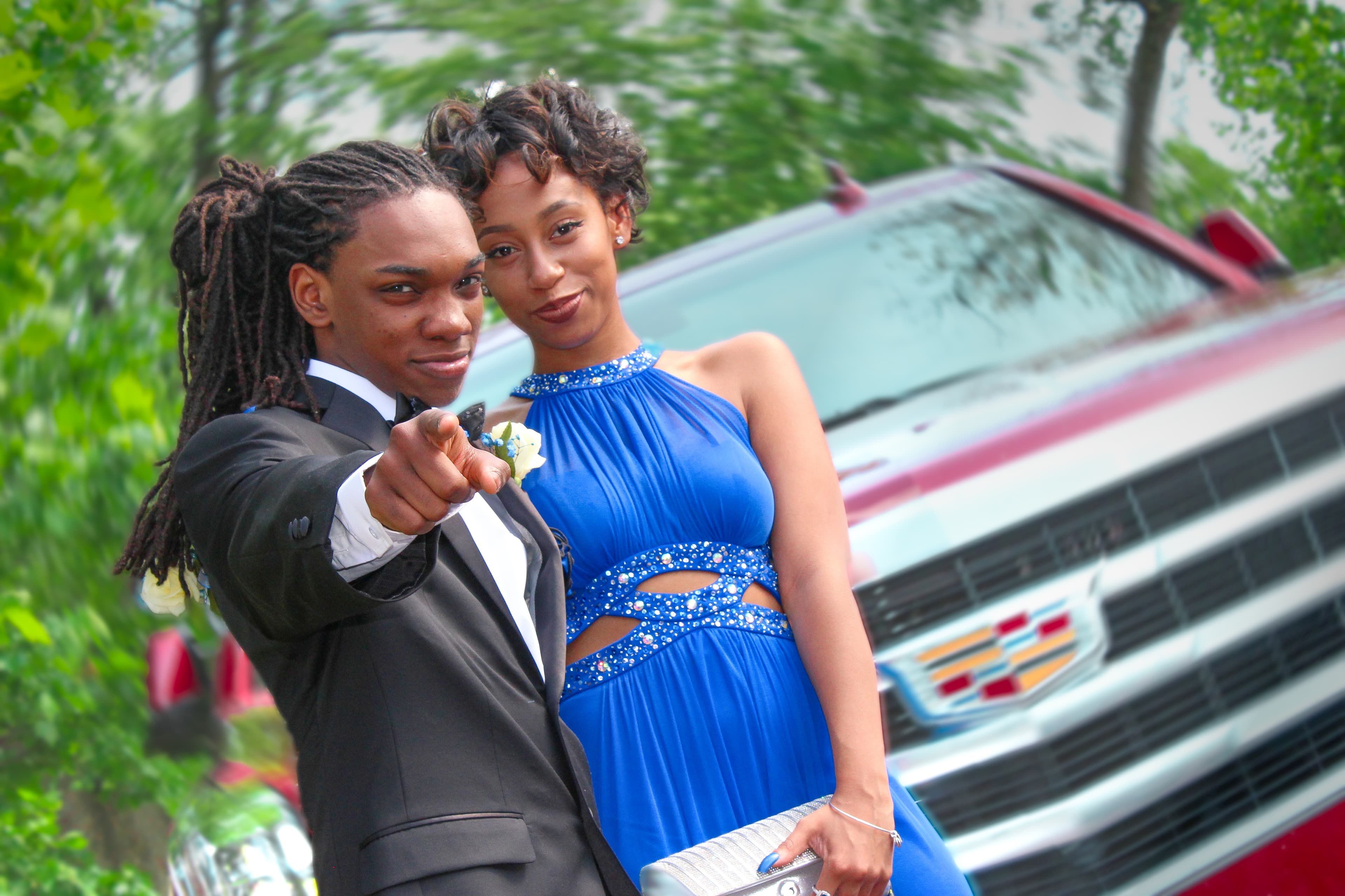 Young couple in formal attire posing outdoors in front of a red Cadillac SUV.