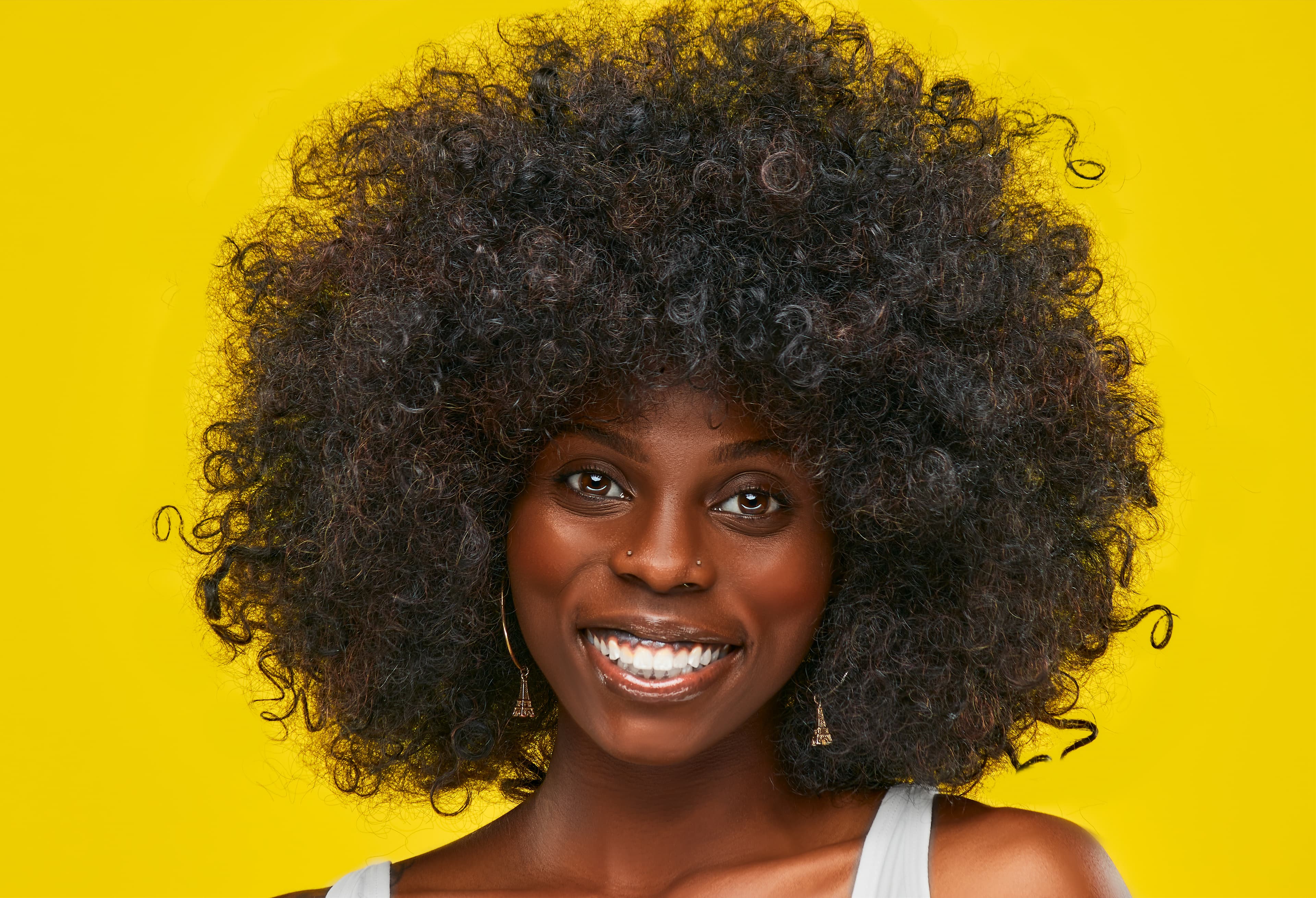 A smiling Black woman with a large, voluminous afro hairstyle against a bright yellow background.
