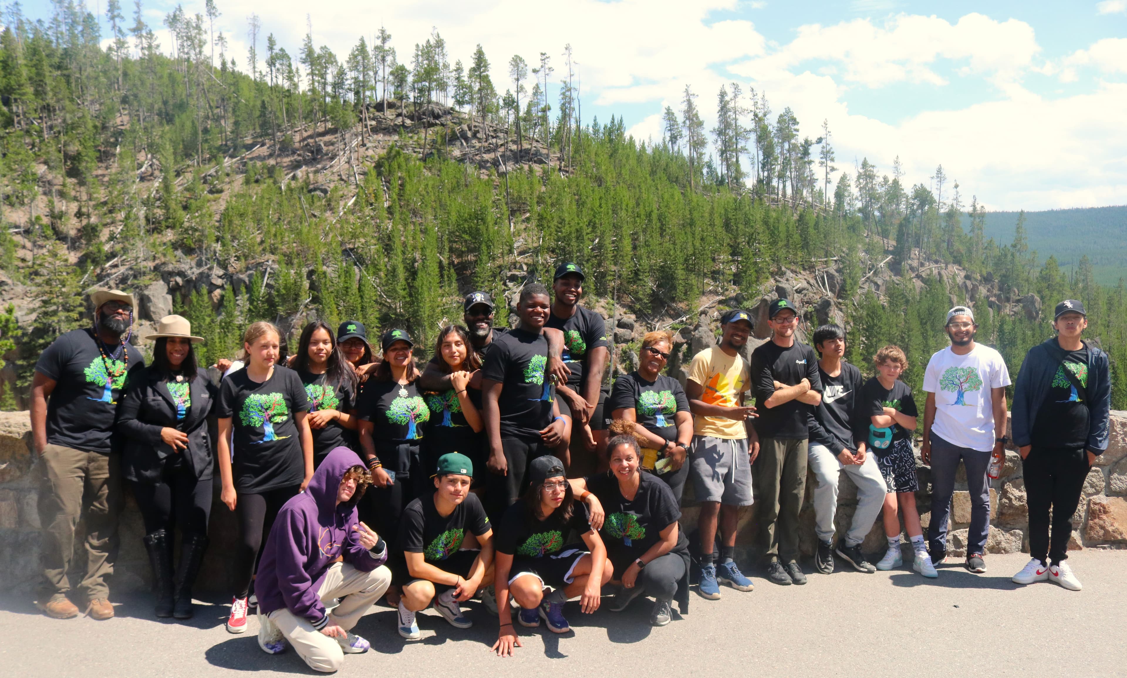 Diverse group of people in matching tree-logo shirts posing before a forested mountain landscape.