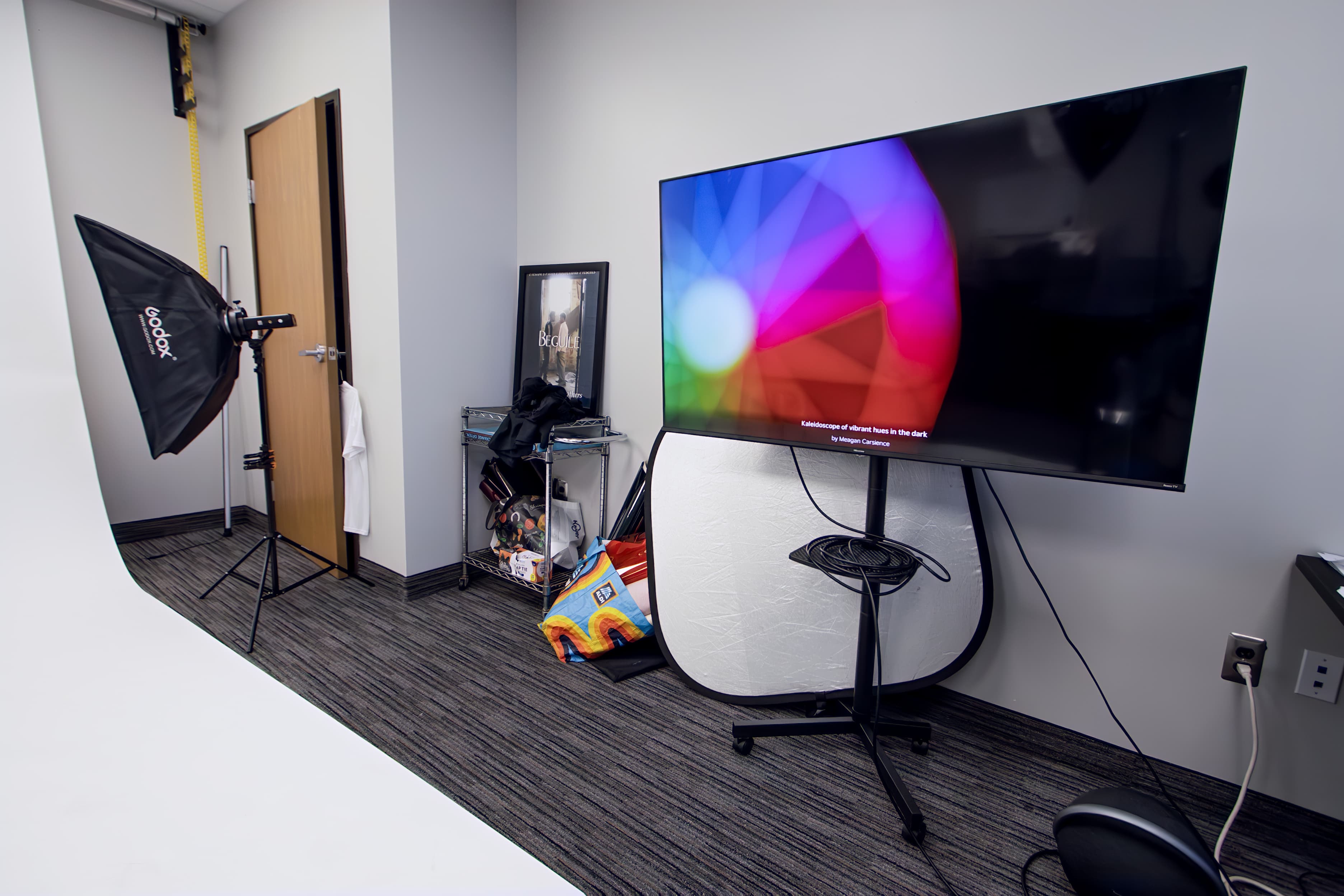 Photography studio featuring a large monitor, softbox light, white reflector, and a white backdrop.