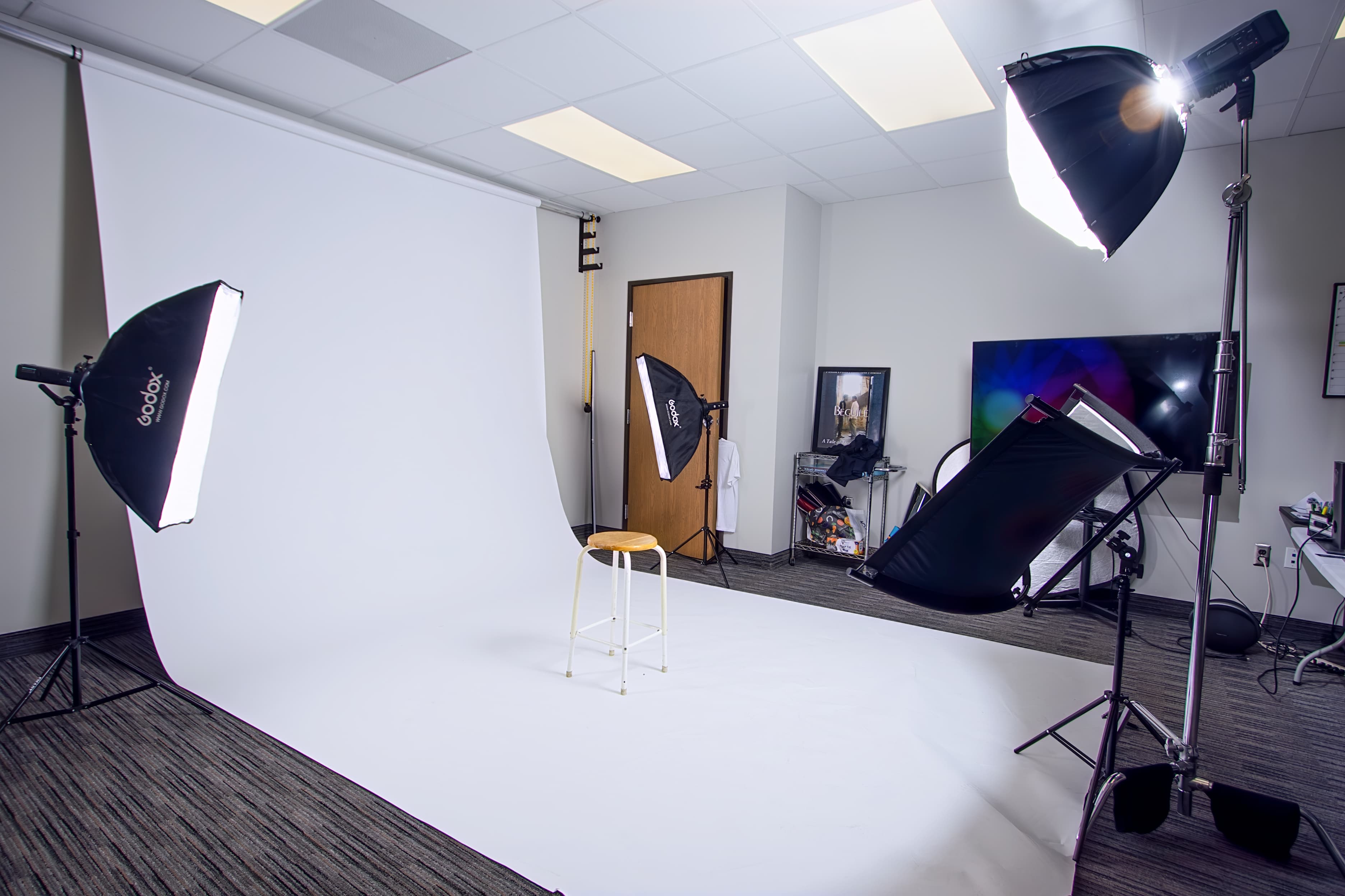 Professional photography studio setup with a white backdrop, softbox lights, and a central wooden stool.